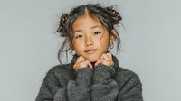 Young Asian girl in cozy gray sweater with hands under chin, messy bun hairstyle on neutral background for lifestyle and fashion content.