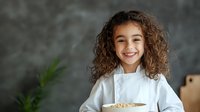 Young mixed race girl with curly hair wearing chef uniform holds bowl of ingredients in professional kitchen setting against dark textured wall, expressing joy and confidence.