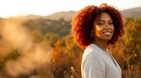 Young African American woman with curly hair smiling outdoors during golden hour sunset with mountains and autumn foliage in background.