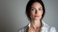Confident businesswoman with natural makeup wearing white shirt and delicate necklace against neutral gray background. Professional portrait for corporate use.