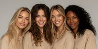 Four diverse women friends smiling together in matching beige shirts against neutral background for teamwork and friendship concepts.