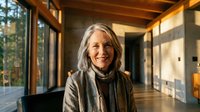Mature woman with long gray hair and red lipstick smiling at camera in modern office hallway with wooden beams and large windows, wearing brown jacket over black top.