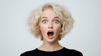 Young Caucasian woman with short blonde curly hair expressing extreme surprise with wide open mouth and eyes against light background. Emotional portrait showing shock and amazement.