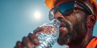 African American construction worker drinking water from plastic bottle on sunny day wearing hard hat and safety glasses for hydration break.