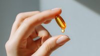 Female hand holding transparent yellow omega-3 fish oil or vitamin D capsule with drop falling on light background. Healthy lifestyle and dietary supplement concept.