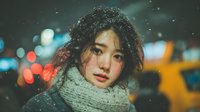 Young Asian woman with curly hair wearing knitted scarf in snowy night city with bokeh lights, creating atmospheric winter portrait.