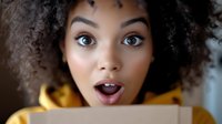 Young African American woman with curly natural hair expressing surprise while opening cardboard package, wearing yellow hoodie against light background.
