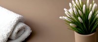Rolled white spa towel and decorative grass plant in ceramic pot against neutral brown background, minimalist bathroom or wellness composition.