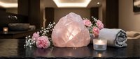 Glowing rose quartz crystal on dark stone surface with pink flowers, candle, and towel in a luxury spa setting.