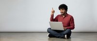 Young African American man in red shirt sitting cross-legged with laptop having eureka moment pointing finger up with idea against white wall background.