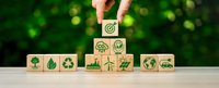 Hand placing wooden cube with target symbol on top of eco-friendly icons including recycling, renewable energy, and sustainability symbols on natural background.