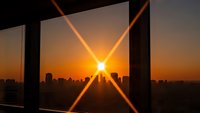 Urban sunrise through modern window frame with dramatic sunburst rays over city skyline. Architectural photography capturing golden hour light and metropolitan landscape.