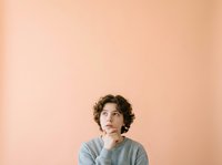 Young man with curly dark hair and olive complexion wearing gray shirt, hand on chin in thoughtful pose against soft peach background for contemplative concepts.