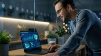 Man in casual blue shirt wearing glasses analyzing financial data and stock market charts on laptop at modern office desk with plants and warm lighting.