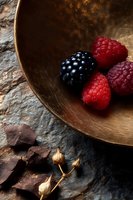 Fresh ripe raspberries and blackberry in copper bowl on dark stone background with chocolate pieces and dried flowers, dramatic food photography.