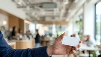 Businessman in blue shirt holding blank white business card in modern office environment with colleagues and natural light for corporate branding.