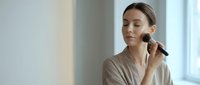 Young Caucasian woman applying makeup with brush in bright modern interior, natural beauty routine and skincare concept for cosmetic advertising.