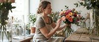 Young Caucasian female florist arranging colorful bouquet in bright flower shop workspace. Professional woman creating floral arrangement for wedding or event.