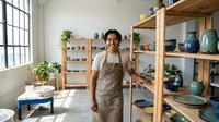Young Asian woman potter smiling in ceramic studio workshop with handmade pottery displayed on wooden shelves and natural light from large windows.