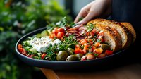 Fresh Mediterranean salad bowl with feta cheese, cherry tomatoes, olives, herbs and grilled bread slices served on dark plate against green background.