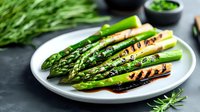 Grilled green asparagus spears with char marks on white ceramic plate, drizzled with balsamic reduction and olive oil, fresh herbs garnish on dark surface.