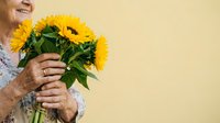 Senior woman with wrinkled hands holding bright yellow sunflowers against pale yellow background, expressing joy and happiness. Suitable for greeting cards, wellness, and lifestyle content.