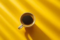 Cup of black coffee on vibrant yellow background with dramatic diagonal shadows creating minimalist composition and modern aesthetic appeal.