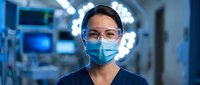 Female surgeon wearing protective mask and glasses in modern operating room with medical equipment and bright surgical lights in background.