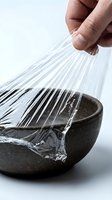Caucasian male hand stretching clear plastic wrap over dark ceramic bowl on white surface, food storage and kitchen preparation concept.