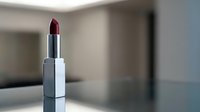 Burgundy lipstick in silver tube standing on reflective surface with blurred modern interior background for beauty and cosmetics advertising.