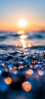 Sunset over ocean waves with golden bokeh lights reflecting on wet pebbles at beach shore, creating magical atmosphere with blue and orange tones.