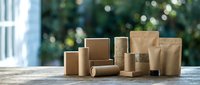 Assorted kraft paper bags and cardboard packaging containers arranged on wooden surface with blurred green foliage background, suitable for e-commerce, retail, and sustainable packaging concepts.