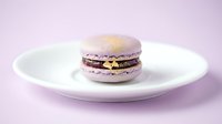 Elegant lavender macaron with gold leaf detail on a white plate, set against a soft purple background, perfect for bakery and food menus.
