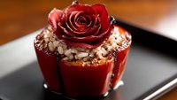 Elegant red rose adorns gourmet dessert with crushed nuts and chocolate drizzle on black plate, dramatic lighting creates luxurious mood for fine dining presentation.