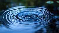 Single water drop creating concentric ripple circles on dark blue water surface with suspended droplet frozen in time, capturing moment of impact.