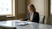 Middle-aged Caucasian woman, professional businesswoman wearing glasses and dark blazer, reviewing documents at marble desk in modern office setting, suitable for corporate, business, and workplace concepts.