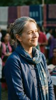 Senior Caucasian woman with gray hair smiling warmly in traditional blue ethnic clothing at outdoor cultural market event with colorful background.