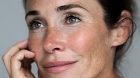 Close-up portrait of middle-aged Caucasian woman with freckles, natural beauty, and expressive blue eyes against neutral gray background.