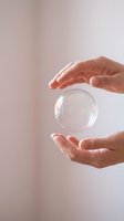 Hands holding transparent glass sphere with iridescent reflections against neutral background, symbolizing care, protection, fragility, and sustainability concepts.