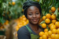 Young African woman in headwrap and overalls smiling while harvesting fresh oranges in citrus grove, agricultural worker during fruit picking season.