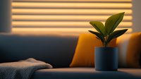 Cozy evening interior with rubber plant in gray pot on sofa, warm sunlight through blinds casting stripes, yellow cushions and textured throw blanket creating relaxing atmosphere.
