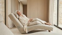 Mature woman with gray hair and light skin tone, wearing beige casual clothing, reclining on a curved upholstered lounge chair in a minimalist interior space with neutral tones and natural light from windows.