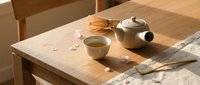 Peaceful tea ceremony setup with ceramic teapot and cup on wooden table with scattered pink petals and bamboo whisk in warm natural lighting.