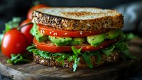 Fresh sandwich with whole grain bread, crisp lettuce, ripe tomato slices and avocado on rustic wooden board, close-up view in natural lighting.