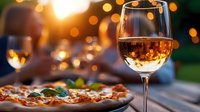 Glass of rose wine and fresh pizza on wooden table outdoors with warm sunset bokeh lights creating romantic atmosphere for summer dining experience.