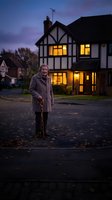 Senior Caucasian man with walking cane standing outside Tudor-style house at twilight with warm glowing windows creating cozy residential evening atmosphere.