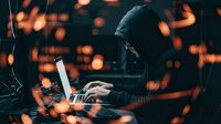 Hooded hacker typing on laptop computer in dark room with glowing orange bokeh lights creating mysterious cybercrime atmosphere for security concepts.
