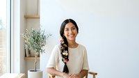 Young woman with long dark hair and South Asian ethnicity wearing casual beige t-shirt, smiling at camera in bright modern interior with potted plant and wooden shelves behind her.