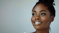 Young African American woman with radiant smile and glowing skin against soft gray background, wearing natural makeup and hair in elegant updo. Professional portrait.