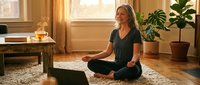 Middle-aged woman of Caucasian ethnicity in lotus position practicing yoga or meditation indoors with laptop, plants, and warm natural lighting for wellness and mindfulness content.